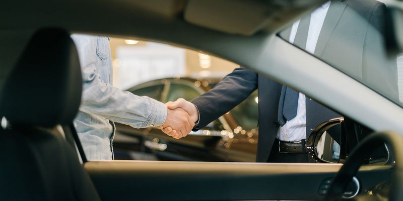 Bilde fra innsiden av en bil hvor man ser to personer som tar hverandre i hendene i en bilforhandler.
