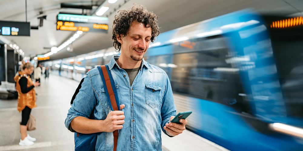 En kille står på perrongen och tittar ner i sin mobil, medan tunnelbanan åker förbi.