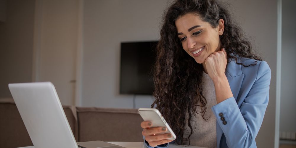 En kvinna med mörkt hår och blå kavaj sitter vid bordet och ler när hon tittar ner i sin mobil. På bordet är en laptop uppfälld och i bakgrunden syns en tv.