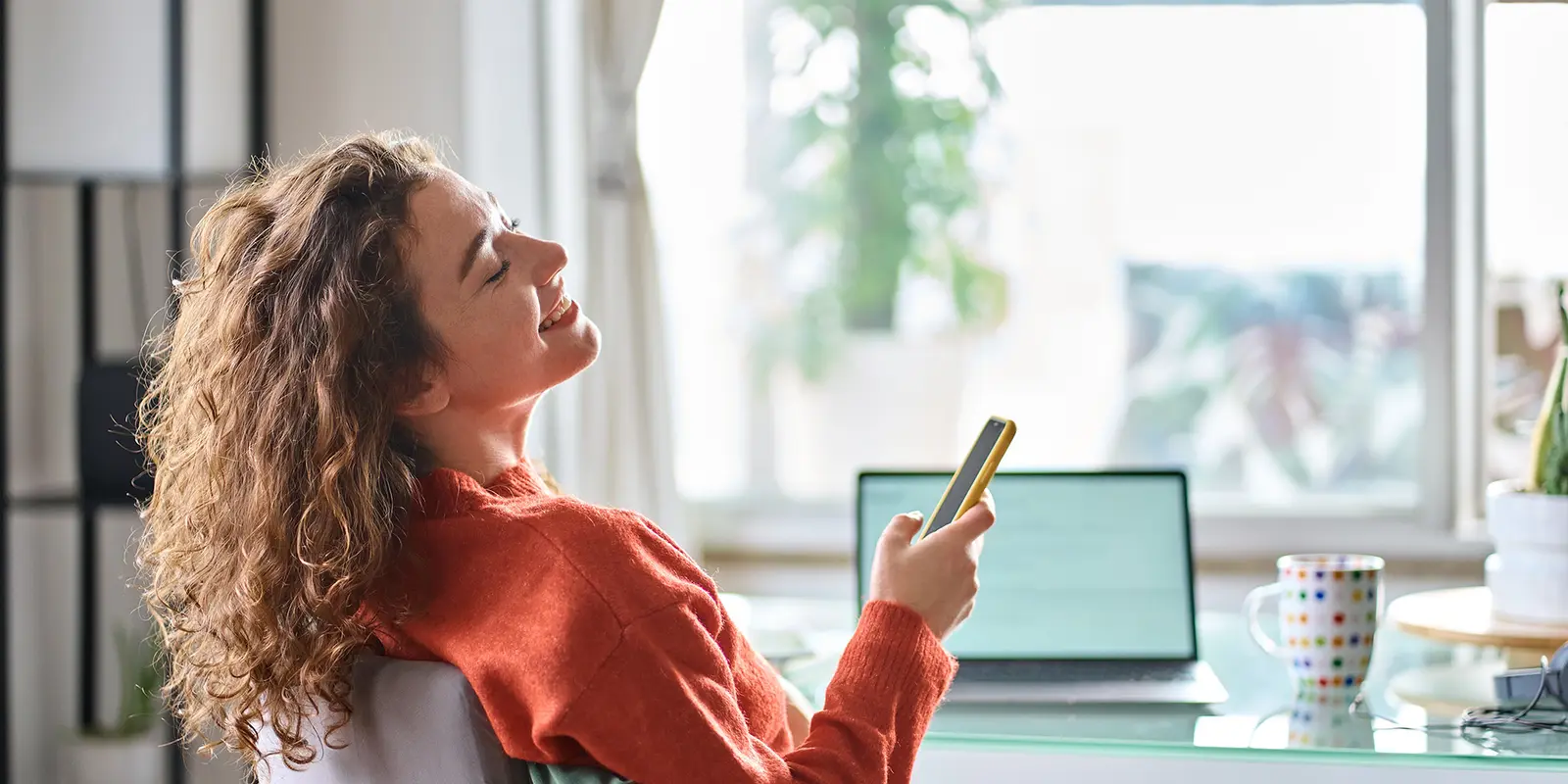 Bilde av en kvinne som smiler mens hun ser opp fra mobilen og pcen.