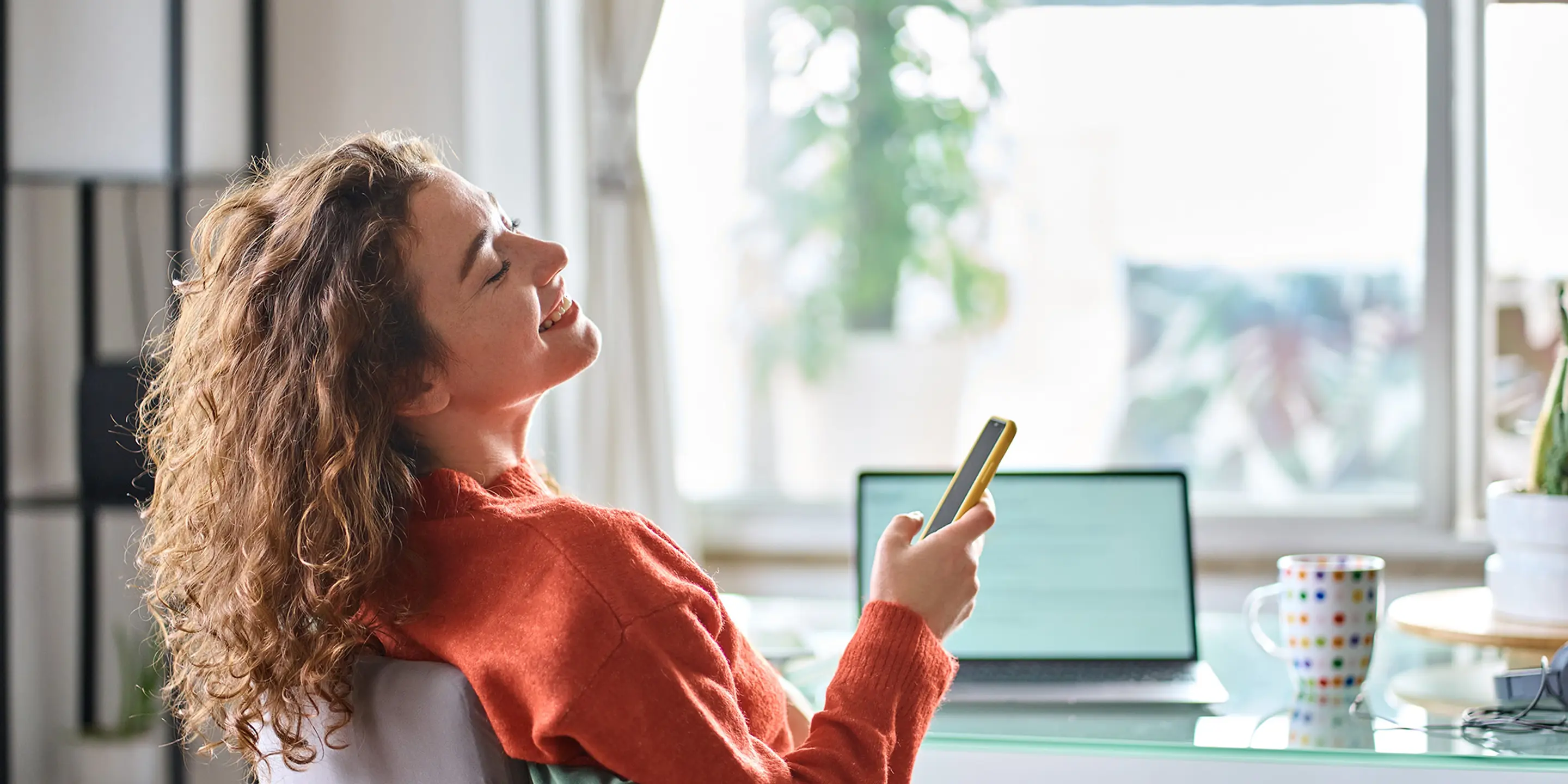 Bilde av en kvinne som smiler mens hun ser opp fra mobilen og pcen.