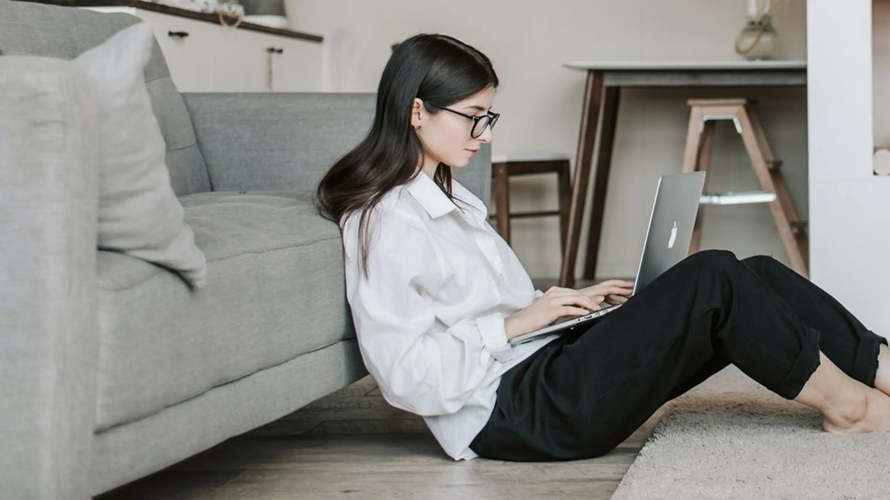 Bilde av en kvinne som sitter på gulvet inntil en sofa og skriver på en laptop.