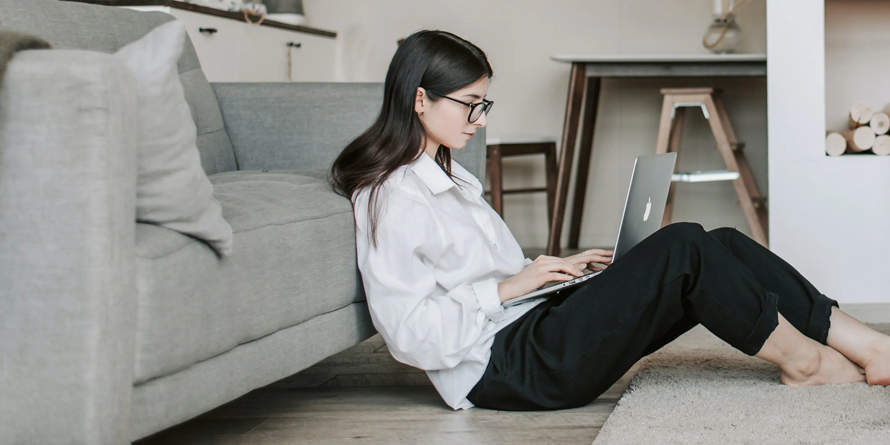 Bilde av en kvinne som sitter på gulvet inntil en sofa og skriver på en laptop.