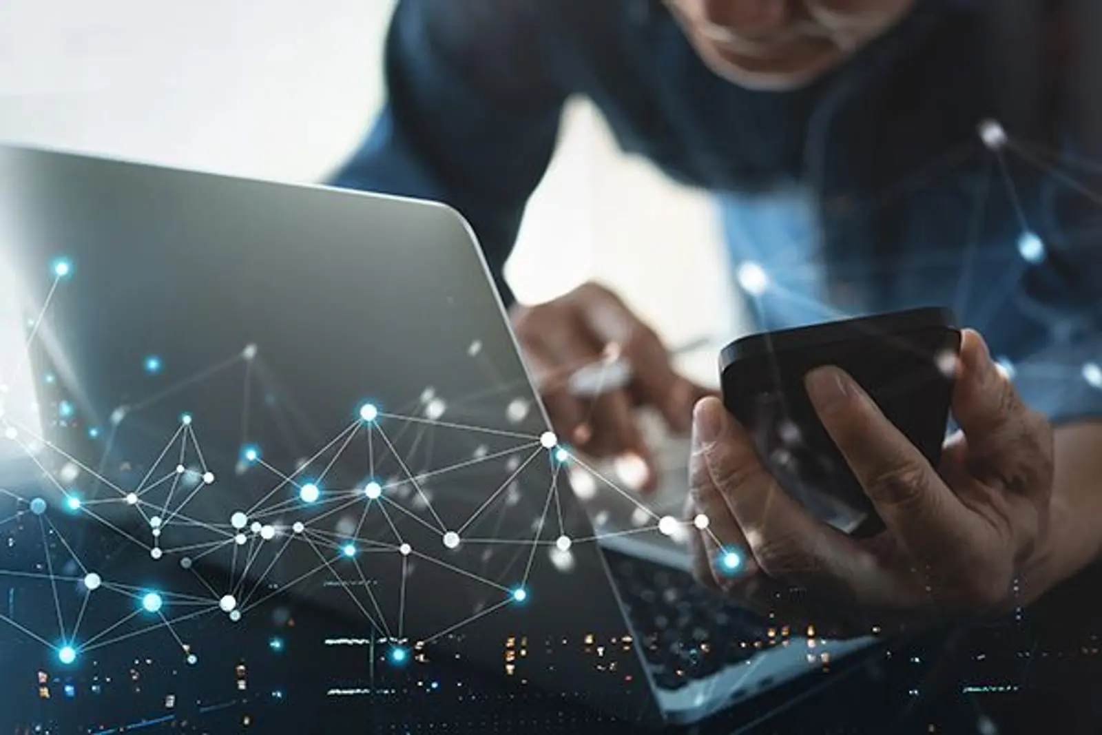 Bilde av en person som står ved en laptop med mobil i hånda