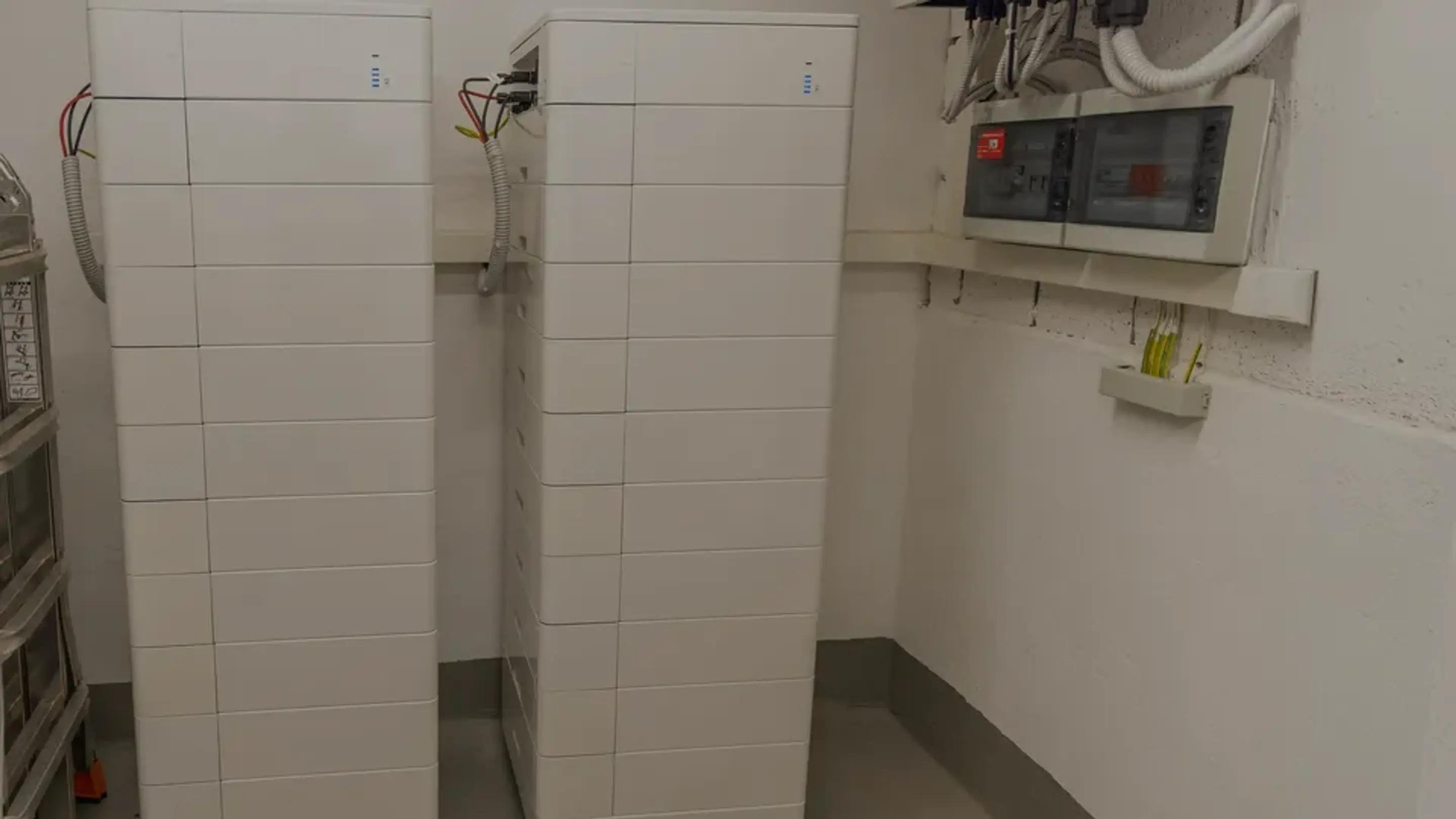 Två höga vita hembatterier som står sida vid sida i ett teknikrum, anslutna till en elcentral på väggen.