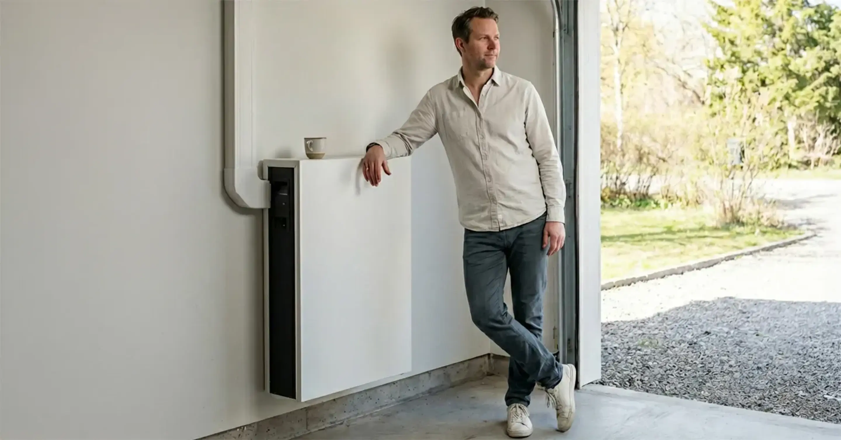En stolt medelålders svensk man som lutar sig mot sitt nyinstallerade hembatteri och ser nöjd ut. Han har ställt en kaffekopp på batteriet.