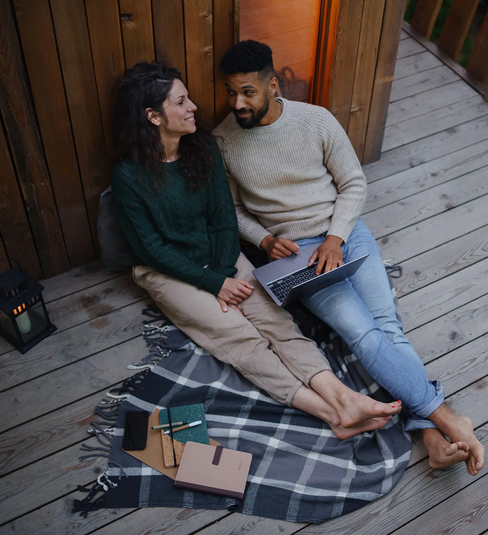 Bilde av et ungt par som sitter på terassen utenfor en hytte og ser på hverandre mens de bruker en laptop.