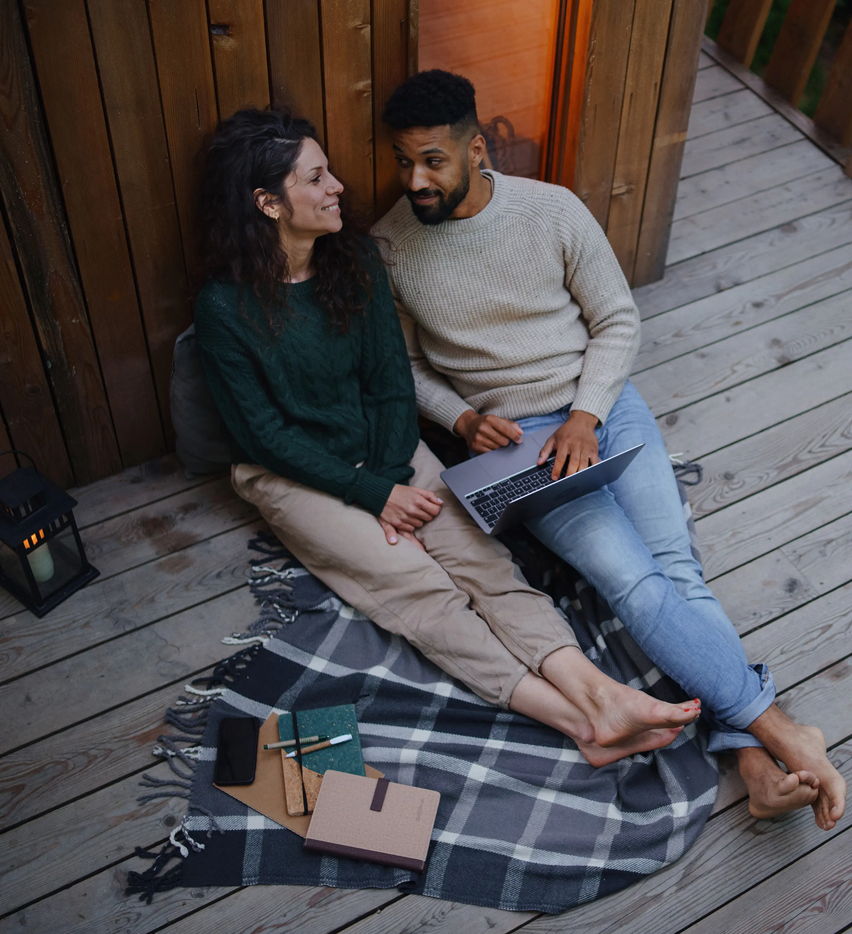 Bilde av et ungt par som sitter på terassen utenfor en hytte og ser på hverandre mens de bruker en laptop.