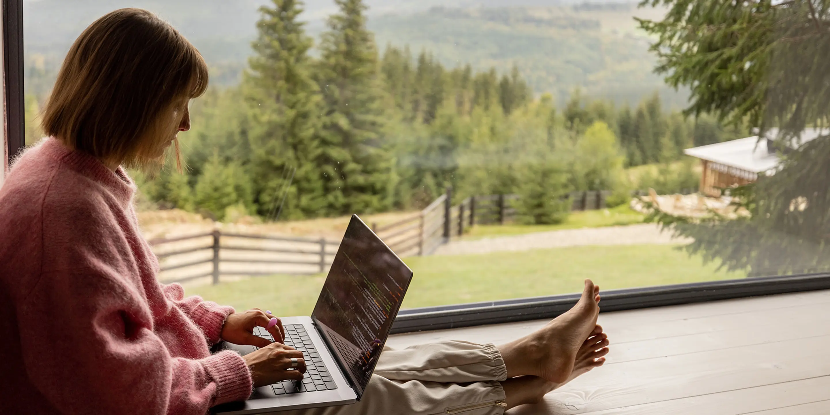 Bilde av en kvinne som sitter ved et stort vindu og skriver på en bærbar pc.