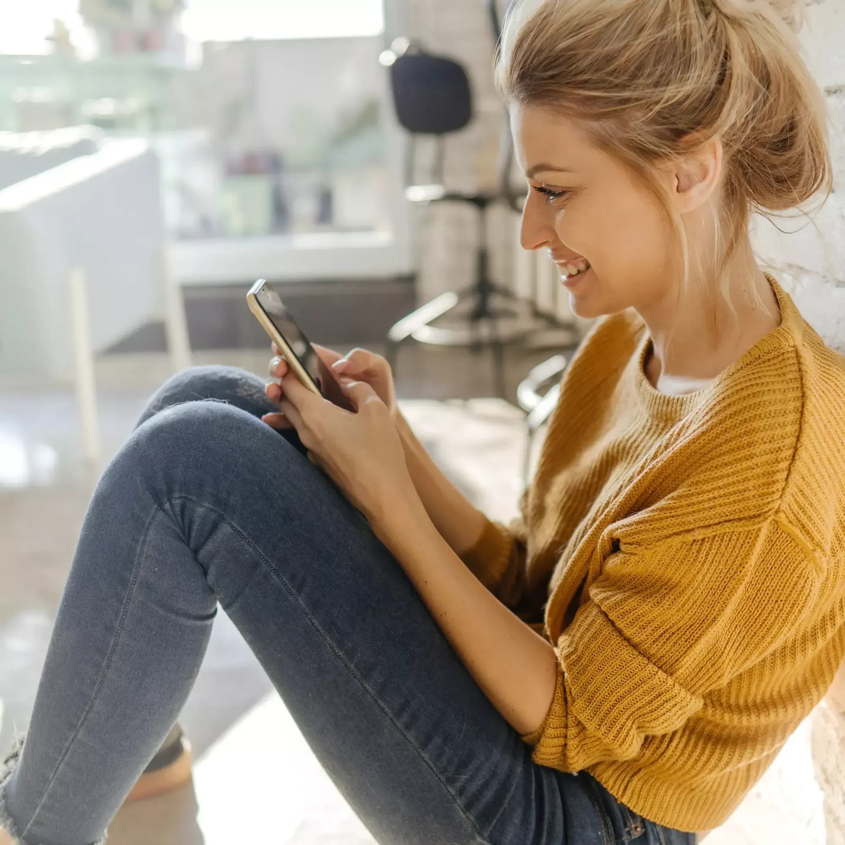 Kvinna i solbelyst vardagsrum skriver med mobilen.