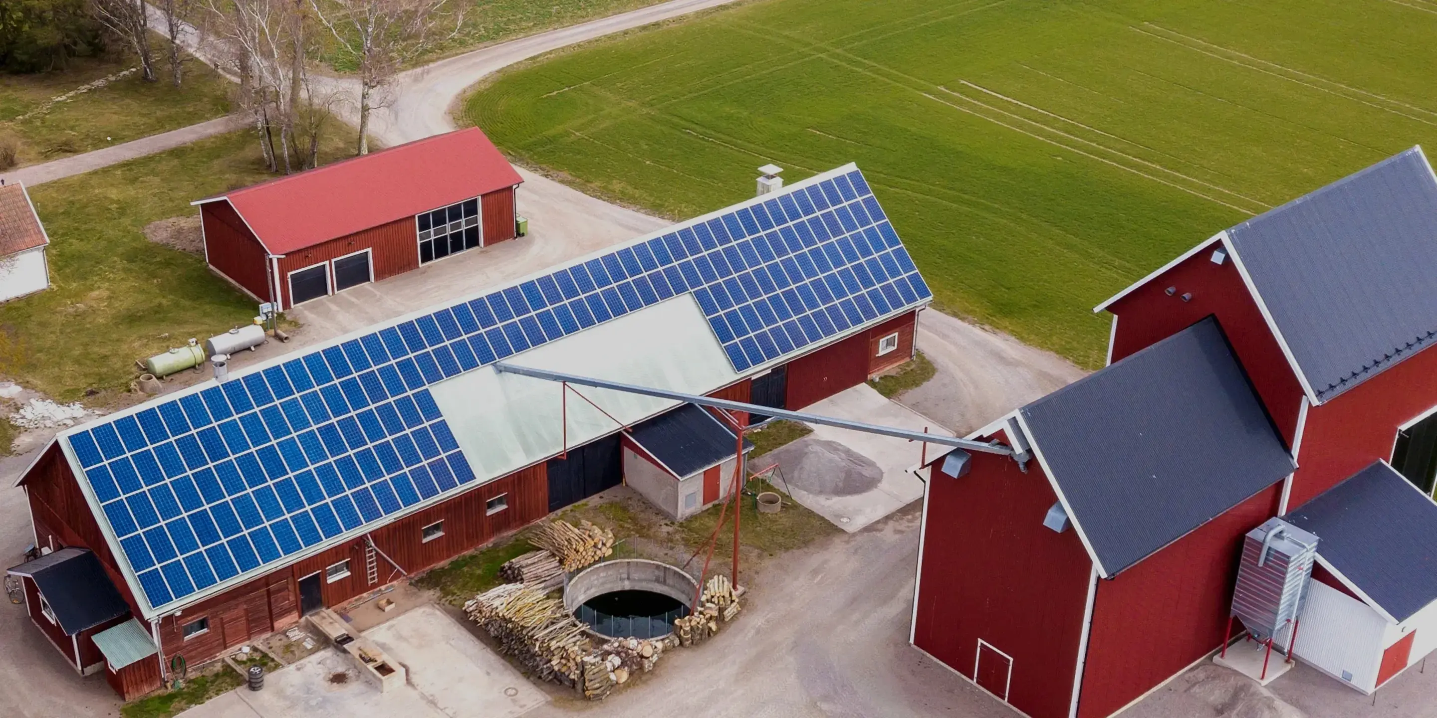 Flygbild över röda gårdsbyggnader med solpaneler på taken omgivna av åkermark.