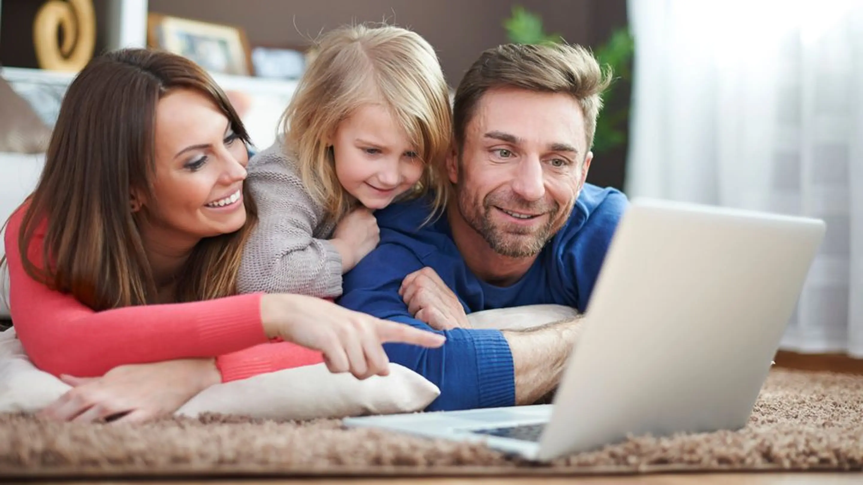 Bilde av et foreldrepar som ligger på gulvet og ser på en laptop med et barn som ligger på ryggen deres og ser.