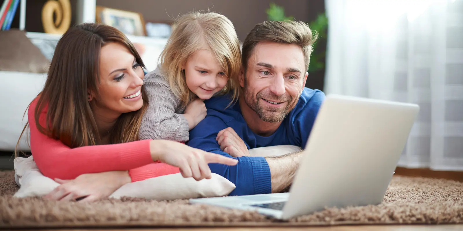 Bilde av et foreldrepar som ligger på gulvet og ser på en laptop med et barn som ligger på ryggen deres og ser.