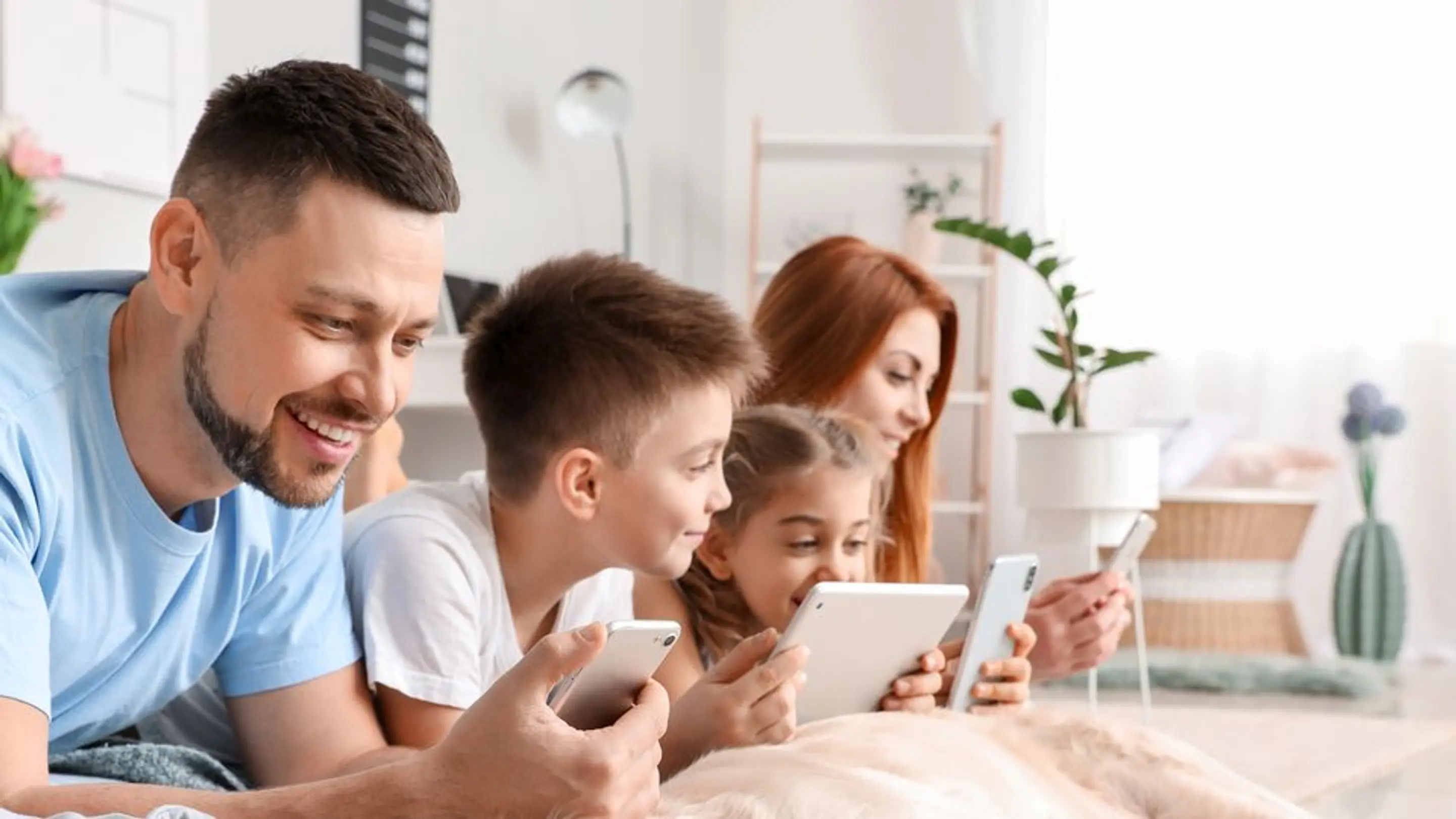 Bilde av en smilende familie som ligger på gulvet og ser på hver sin skjerm.