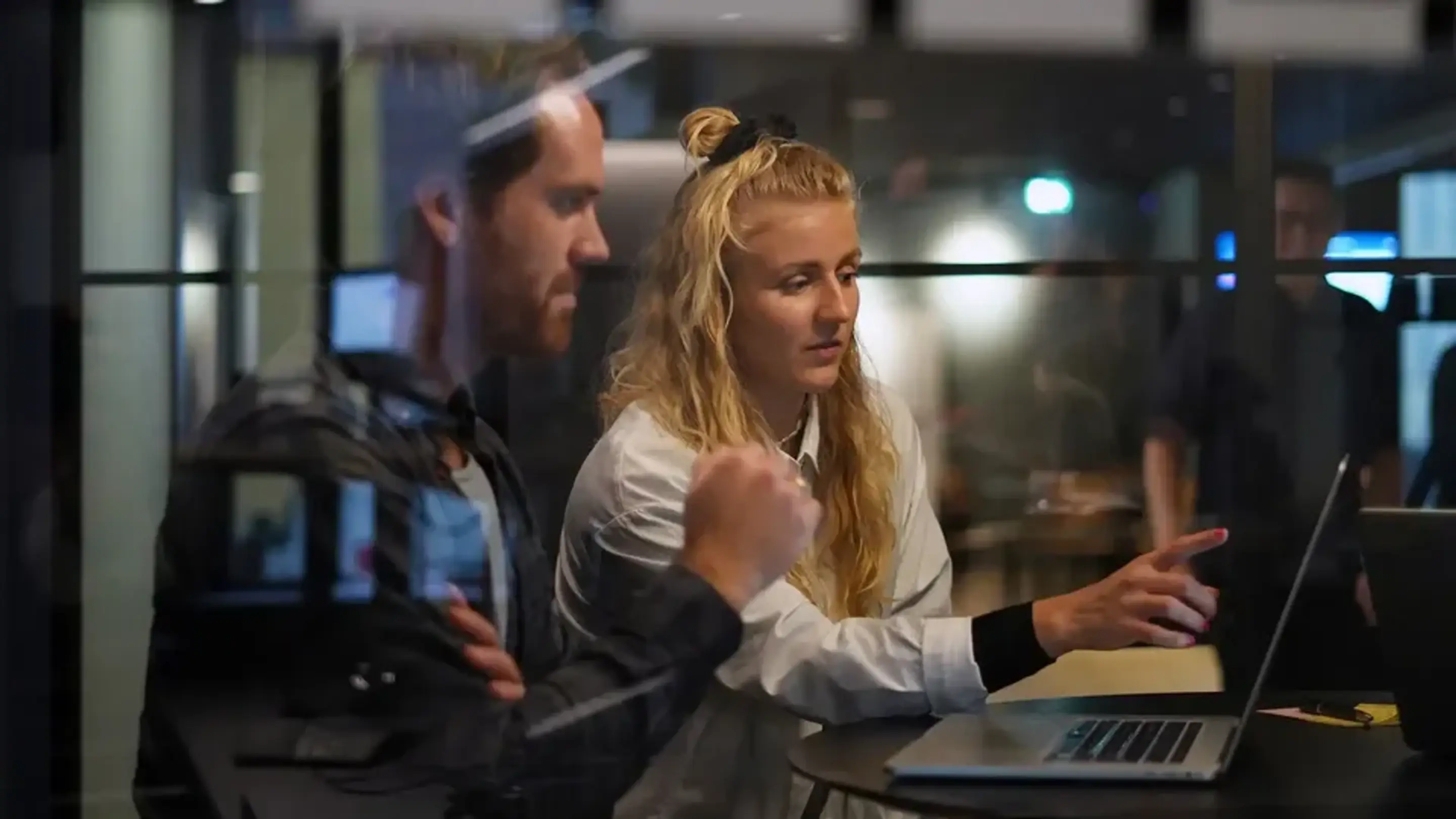 Två kollegor samarbetar vid en laptop i ett modernt kontor och diskuterar något på skärmen.