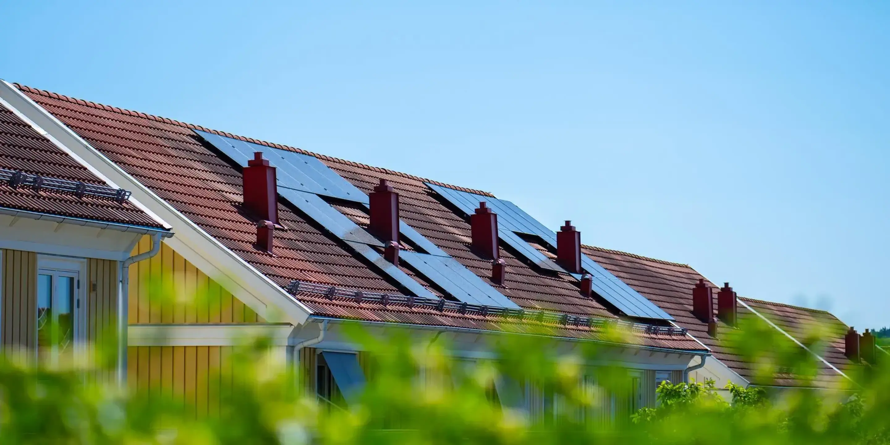 Solens strålar träffar solpanelerna på ett villatak, med somrig grönska i förgrunden.