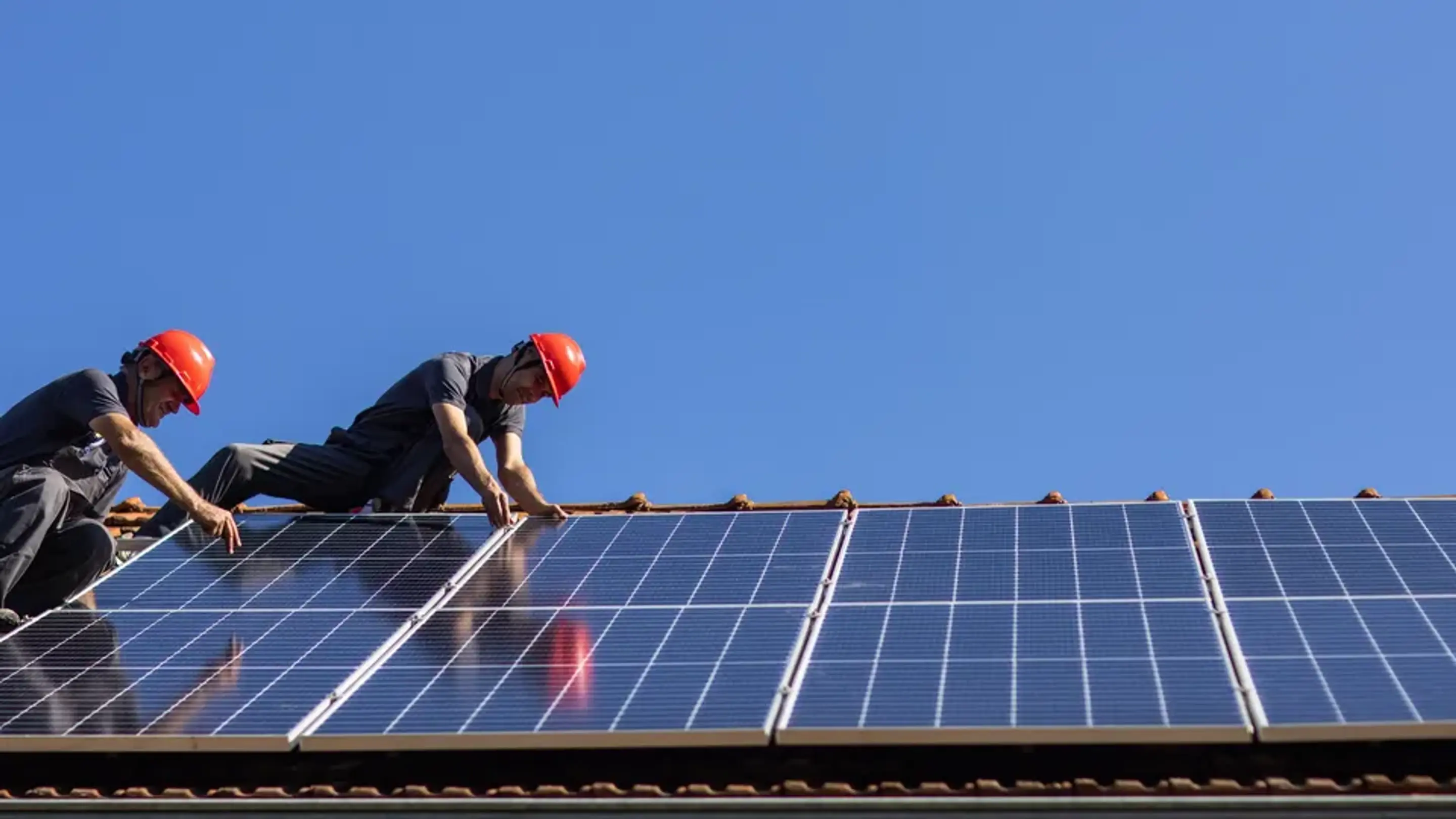 Två installatörer monterar solceller på ett tegeltak i skinande sol.