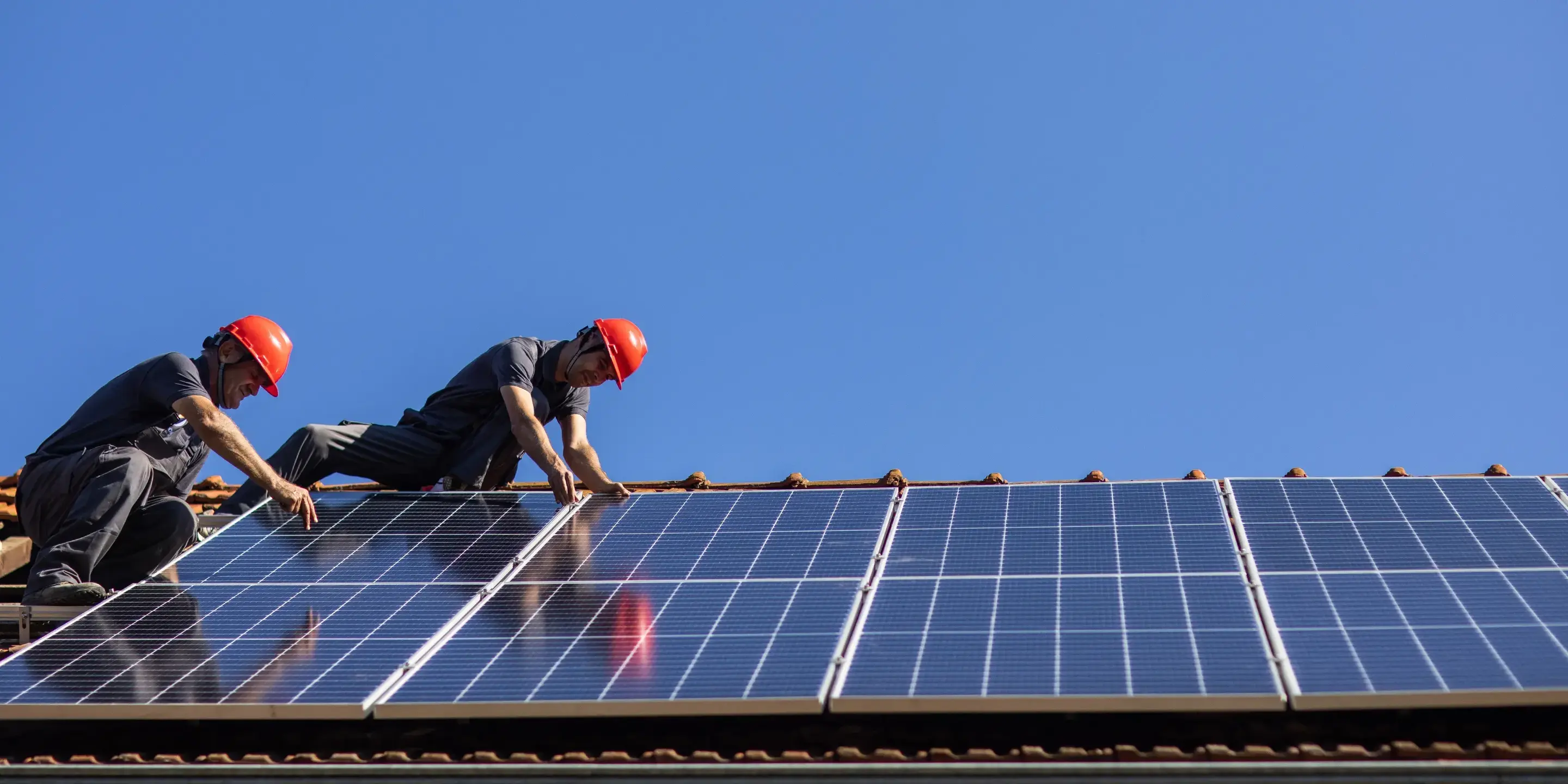 Två installatörer monterar solceller på ett tegeltak i skinande sol.