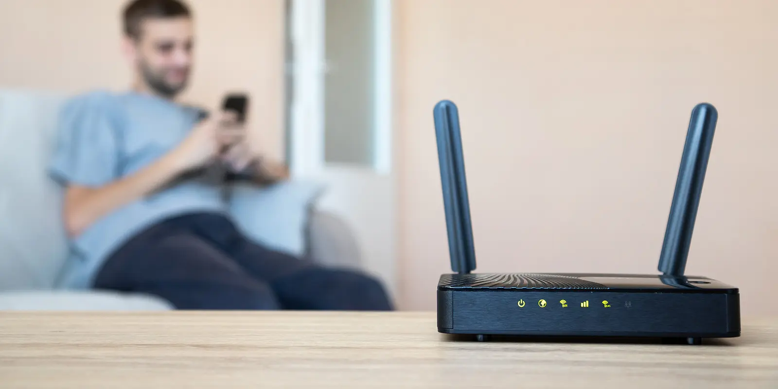 Bilde av en router som står i forgrunnen mens en person sitter på mobilen i en sofa i bakgrunnen.