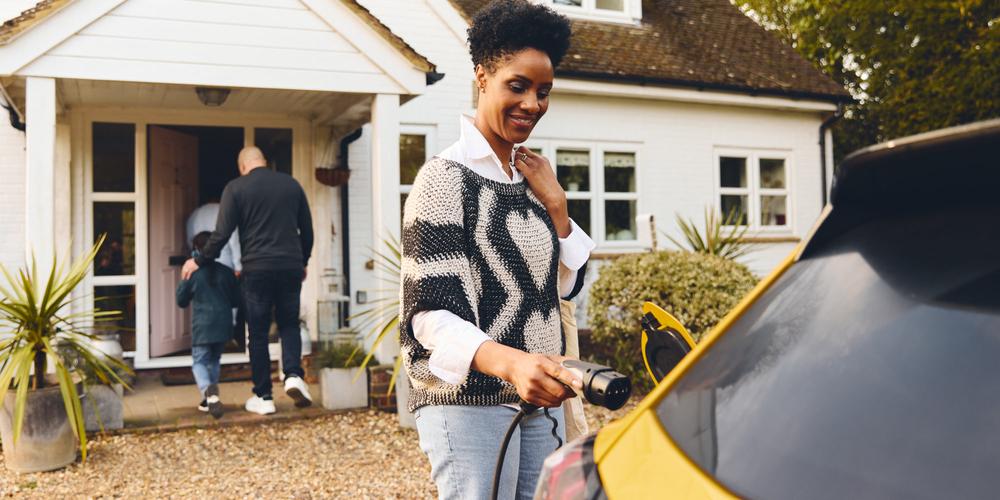 Bilde av en kvinne som plugger i en elbillader mens familien går inn i huset bak henne.