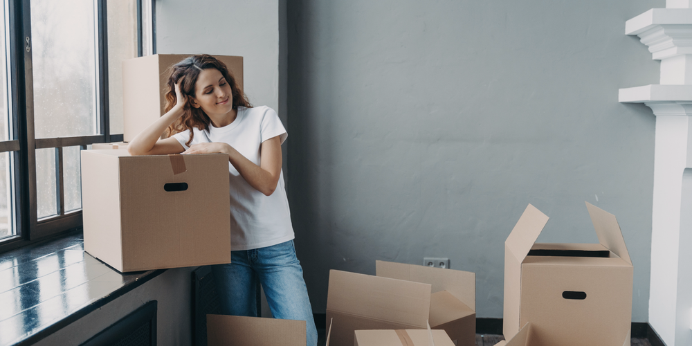 Kvinna står med flyttkartonger i ett rum med flera ouppackade lådor vid en flytt.
