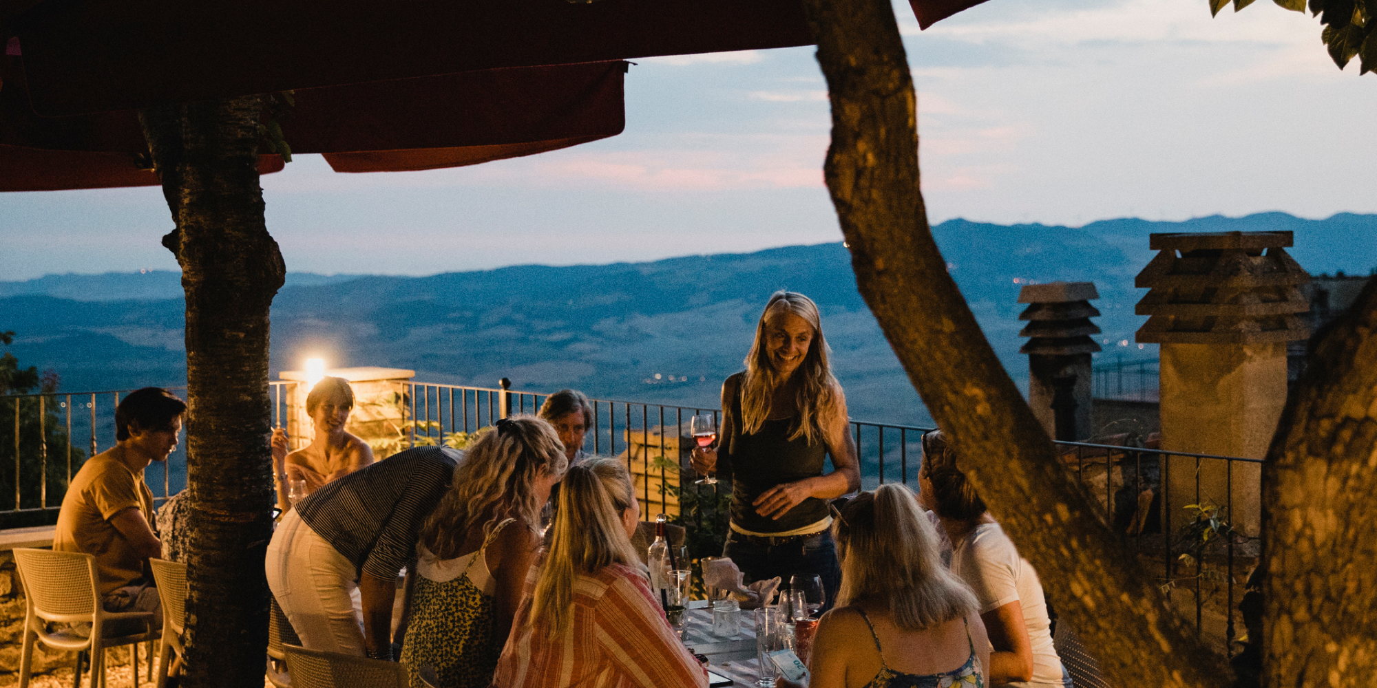 En grupp människor i olika åldrar som äter middag tillsammans på en terrass med utsikt över det italienska landskapet i kvällsljus.