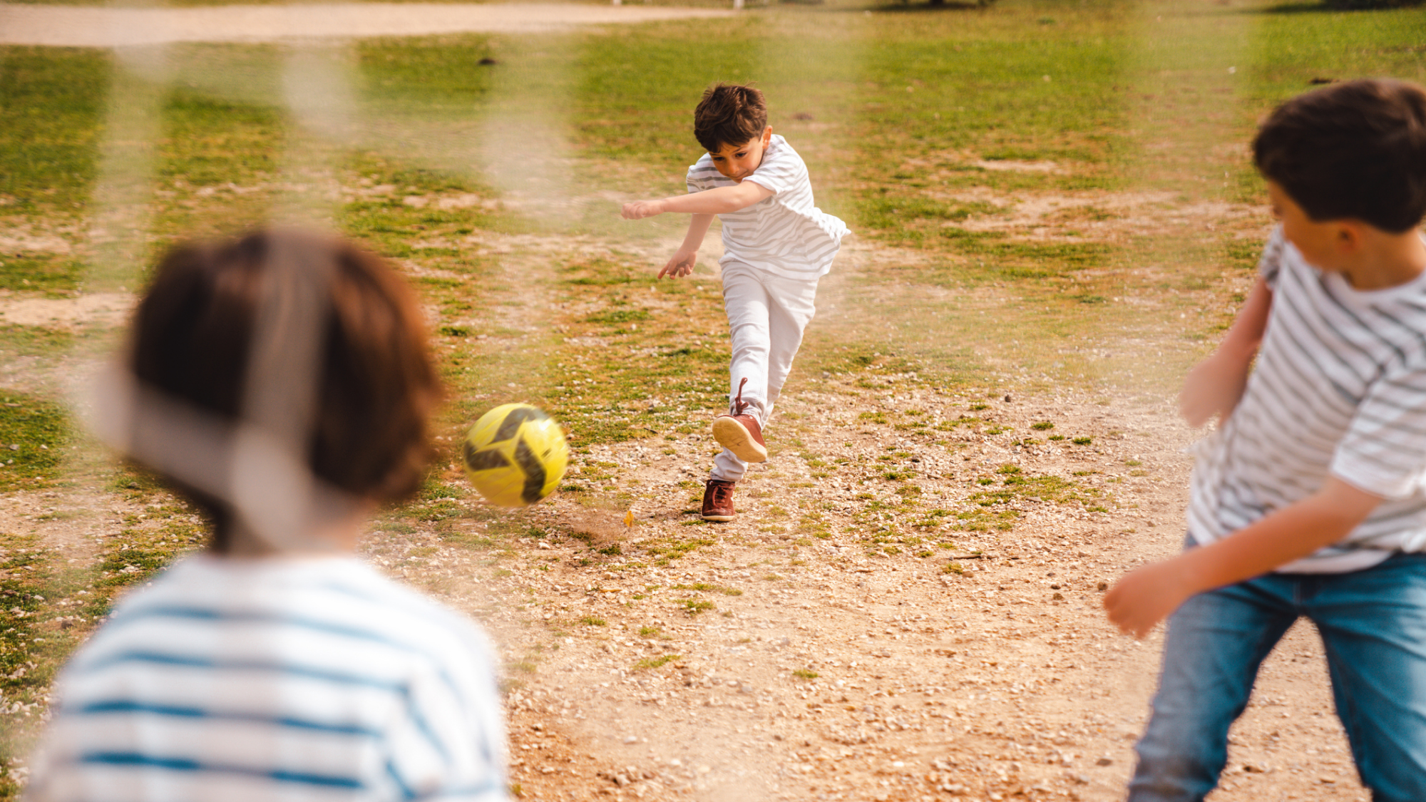Tre pojkar spelar fotboll på en gräsplan. Ena pojken sparkar en gul fotboll mot mål.