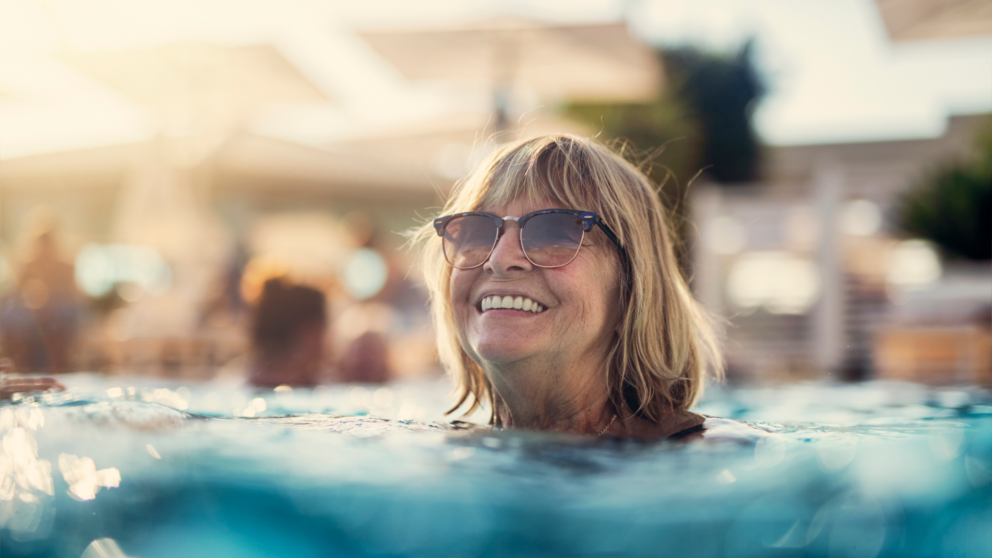En äldre blond kvinna med solglasögon ler i en utomhuspool med vatten upp till axlarna.