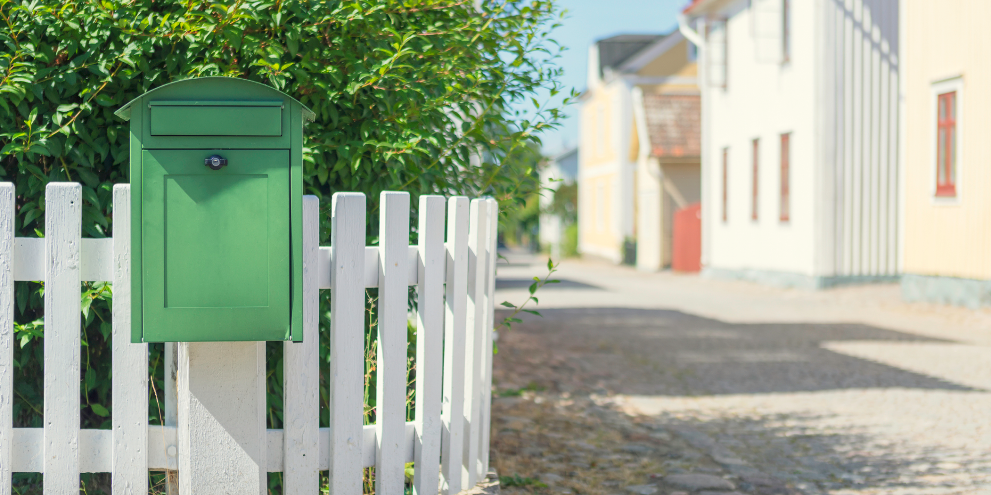 Grön postlåda framför vitt staket i bostadsområde.