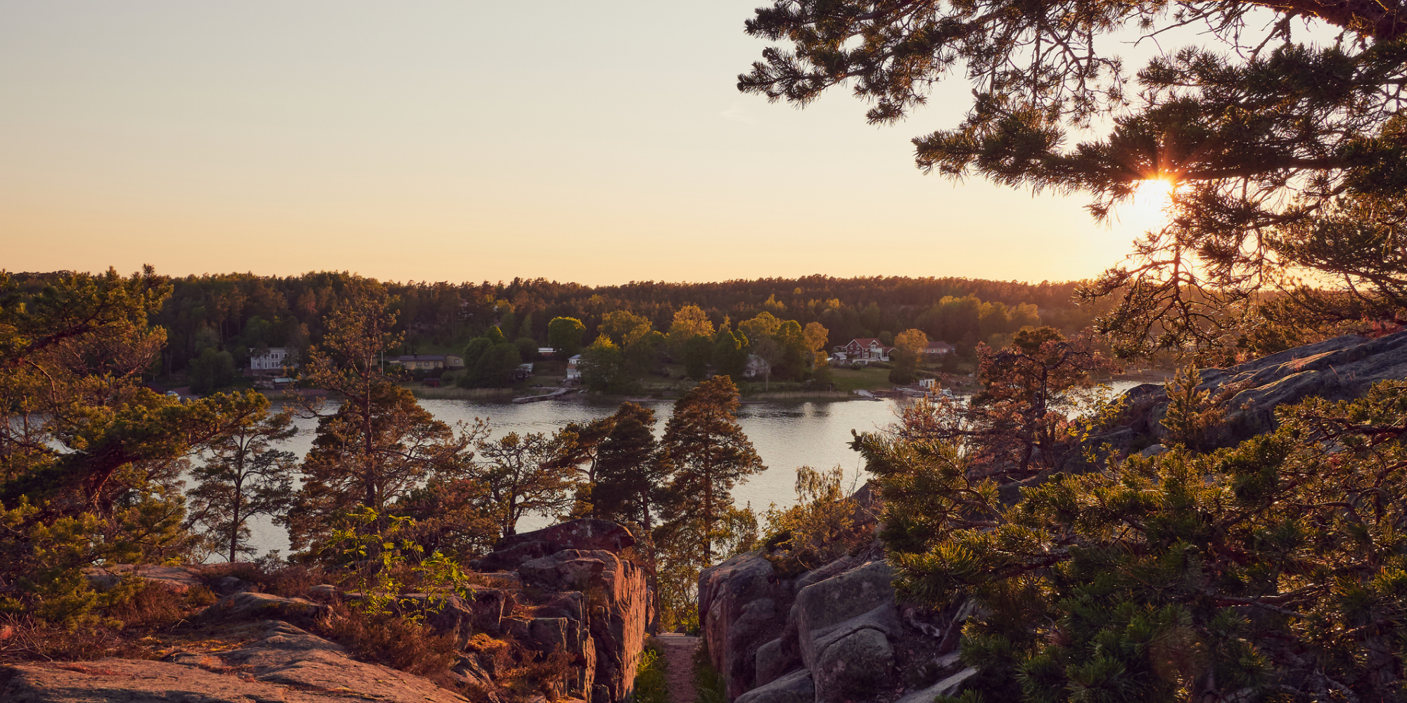 Solnedgång över skärgårdslandskap med klippor, tallar och stilla vatten sett från höjd.