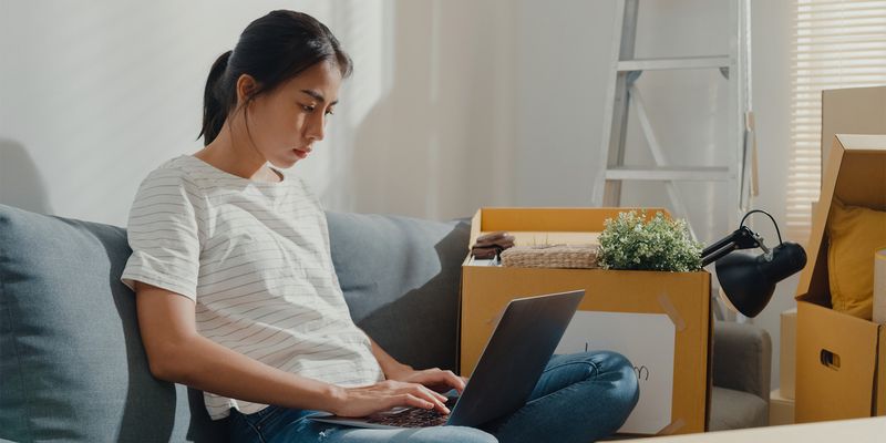 Bilde av kvinne som sitter i en sofa omringet av flytteesker i en sofa mens hun leter opp gode tilbud på flyttehjelp på en laptop.