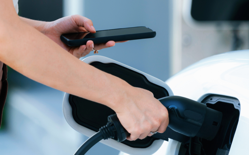 Person ansluter laddkabel till elbil och håller en mobiltelefon i handen.