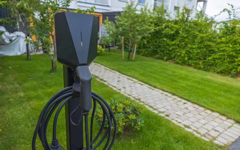 Fristående laddstolpe med laddkabel placerad i trädgård vid bostad.