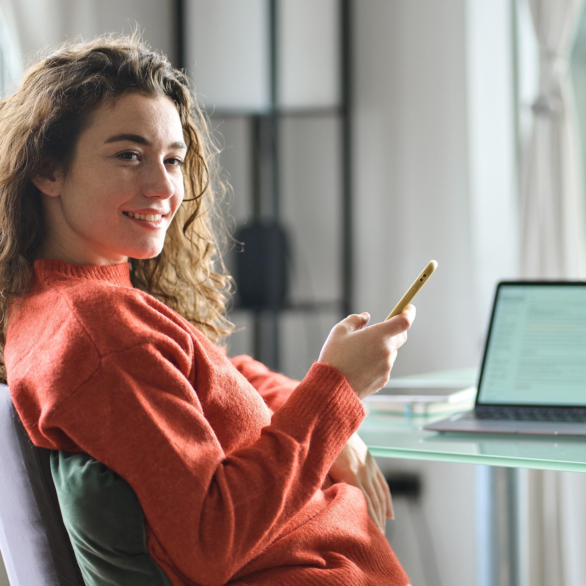 Bilde av en ung kvinne som sitter ved et bord med pc og scroller på mobilen mens hun ser til siden.