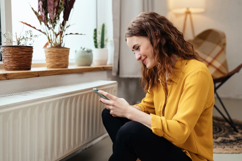 Bilde av en ung kvinne på mobiltelefon som sitter foran en radiator