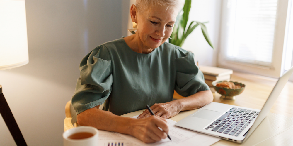 Kvinna med kort ljust hår och grön blus skriver på papper vid köksbordet.
