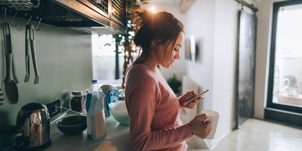 Ung kvinna dricker kaffe i modernt kök och tittar på mobilen medan solen går ner utanför fönstret.