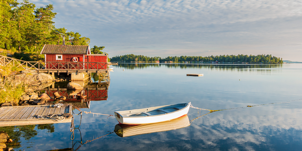 Liten röd stuga vid sjökanten med båt och spegelblankt vatten en klar sommardag i Sverige.
