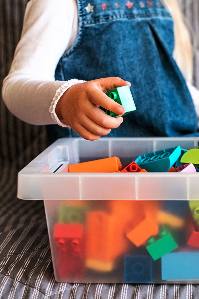 Et barn sitter på en sofa og legger en duplokloss i en stor plastboks full av duplo