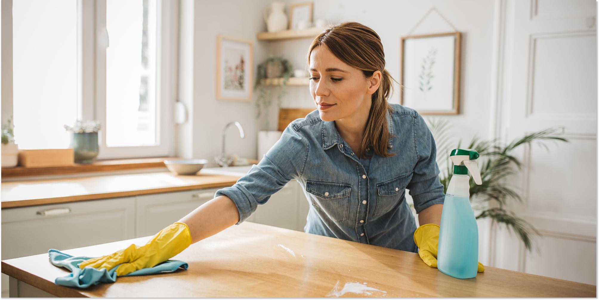 En kvinna med gummihandskar torkar av ytor i köket med en trasa och rengöringsspray, under en hemstädning.